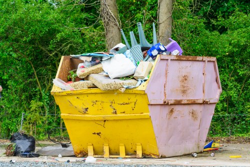 A large, yellow and pink industrial skip overflowing with various types of rubbish, including plastic containers, padded fabric items, cardboard, and miscellaneous waste, positioned on a gravel surface in an outdoor area near a background of green trees and bushes. Some black rubbish bags are placed on the ground nearby. The skip appears weathered, with visible dirt and rust, and is set against a natural, leafy environment, suggesting a site where waste collection or clearing services possibly involve firms like Office Clearance Hampstead Heath operating near the Hampstead Heath area.