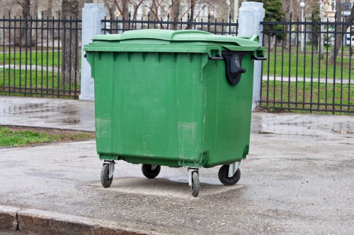 A green plastic wheeled rubbish bin with a closed lid is situated on a paved pathway in an outdoor setting. The bin's surface is smooth with ribbed sides, and it has a black handle on the front for opening the lid. It is positioned against a metal fence with vertical bars and concrete posts, which encloses a grassy area lined with leafless trees, suggesting a park or community space. The ground nearby appears slightly wet, indicating recent rain, and the bin is located near a pavement or sidewalk, typical of urban or suburban environments in the Hampstead Heath area. The scene is evenly lit under natural daylight, emphasizing the utilitarian purpose of the bin for waste disposal and rubbish removal services in the local community, as provided by Office Clearance Hampstead Heath.
