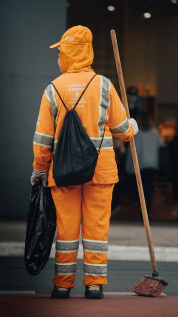 A blue recycling or waste collection cart is positioned on a paved sidewalk next to a tree, with its lid open revealing cleaning tools such as a mop and broom leaning inside. The cart has two large wh