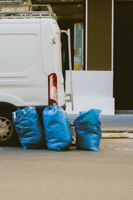 Two large black plastic rubbish bags, visibly filled and tied at the top, are placed on the pavement near the curb in an outdoor setting. The bags appear to contain domestic waste, with some material 