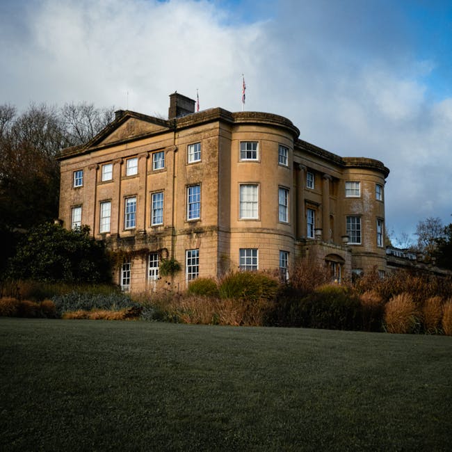 A large, historic stone building with a rounded corner section and multiple rectangular windows, some with white frames, set into a well-maintained facade. The building's exterior features a beige, we